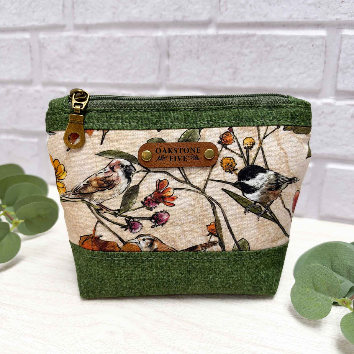 A small zippered pouch with a green vines fabric trim and base, featuring a central printed panel of birds and flowers on a beige background, with a brown faux leather Oakstone Five brand patch on the front.