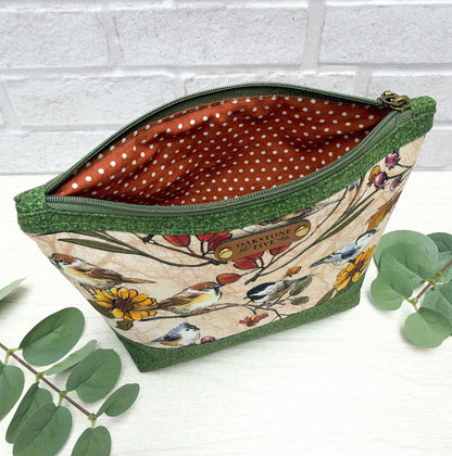 A zippered cosmetic pouch with a green fabric trim and a beige floral-and-bird print exterior, shown open with a rust-orange polka-dot interior. The pouch sits on a white surface with eucalyptus leaves and a white brick wall in the background.
