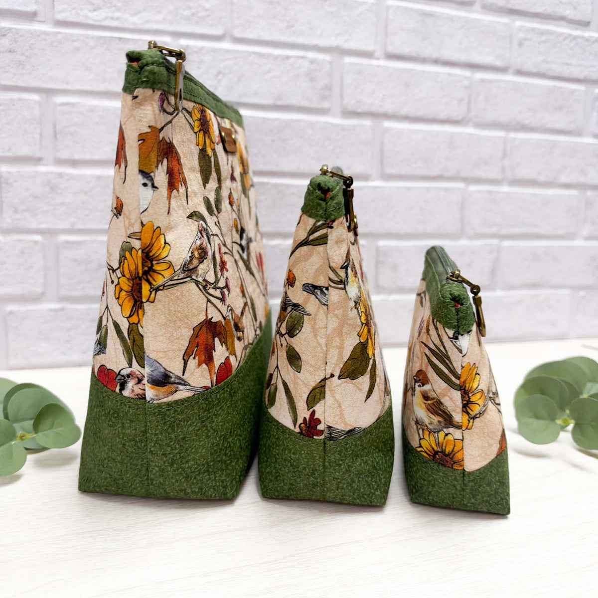 Three fabric zippered pouches in graduated sizes, featuring a beige and green floral and bird print with solid green bases, arranged on a white surface against a white brick wall background.
