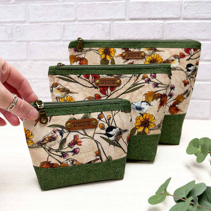 Three zippered cosmetic pouches with a nature bird and floral print, arranged on a white surface against a white brick wall. A hand is holding the smallest pouch on the left, and green leaves are visible in the bottom right corner.