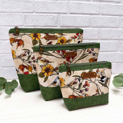 Three fabric zippered pouches in a nested set, featuring a beige background with a colorful bird and floral pattern, dark green textured top and bottom panels, and brown faux leather brand patches. The pouches are styled on a white surface with eucalyptus leaves against a white brick wall background.