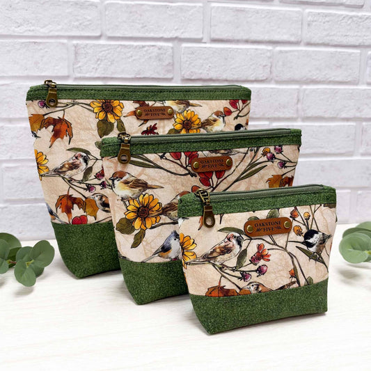 Three fabric zippered pouches in a nested set, featuring a beige background with a colorful bird and floral pattern, dark green textured top and bottom panels, and brown faux leather brand patches. The pouches are styled on a white surface with eucalyptus leaves against a white brick wall background.