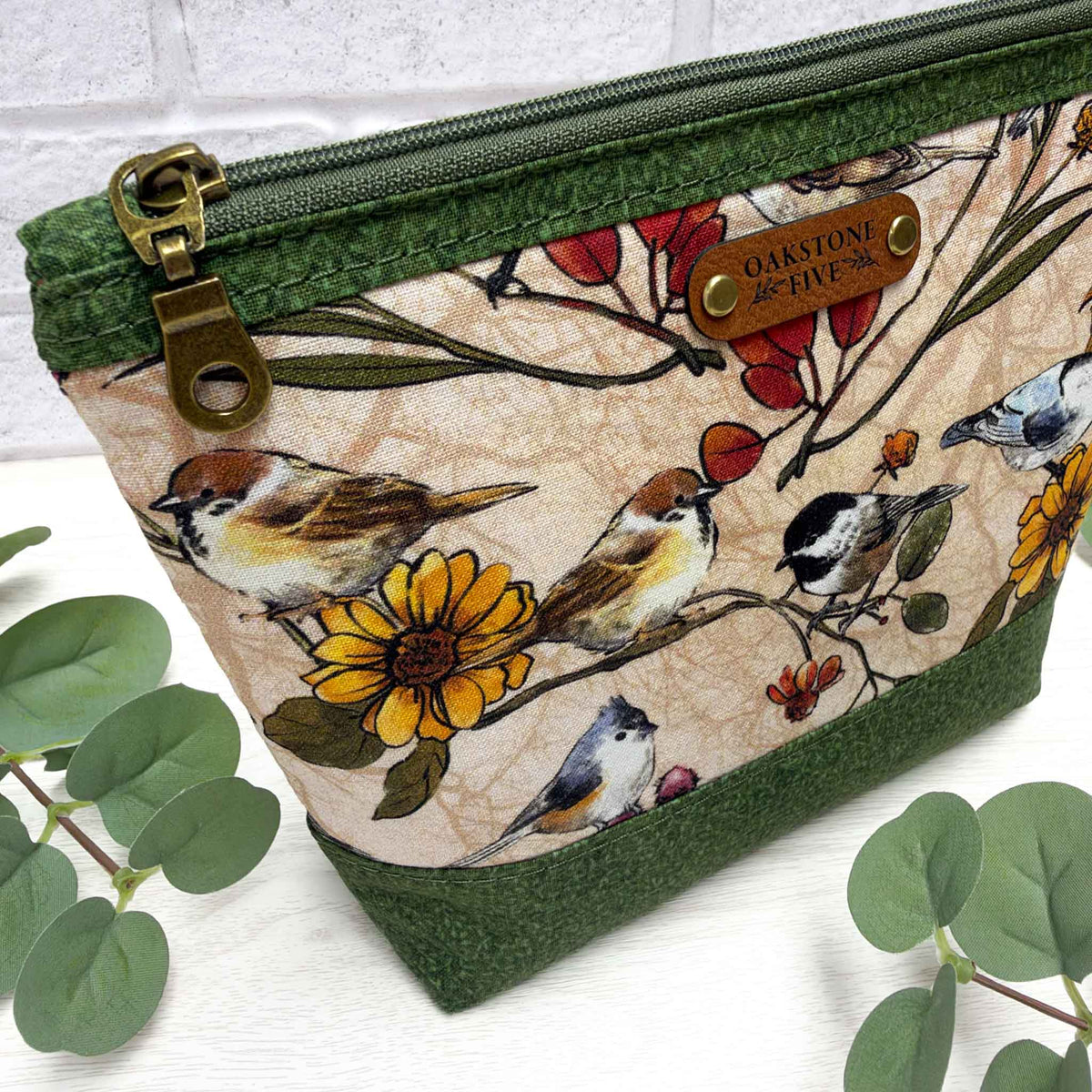 A fabric zip pouch with a green vines bottom and zipper trim, featuring a printed design of birds, flowers, and leaves on a beige background. It has a brass-tone zipper pull and a brown faux leather brand patch.