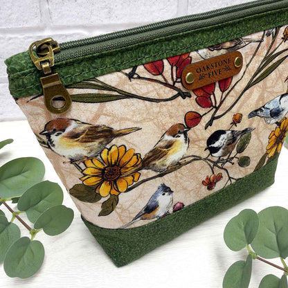A fabric zip pouch with a green vines bottom and zipper trim, featuring a printed design of birds, flowers, and leaves on a beige background. It has a brass-tone zipper pull and a brown faux leather brand patch.