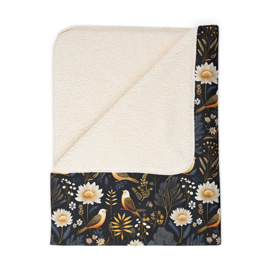 A folded, nature-inspired sherpa blanket featuring a dark background with a bird and botanical motif, on a white background