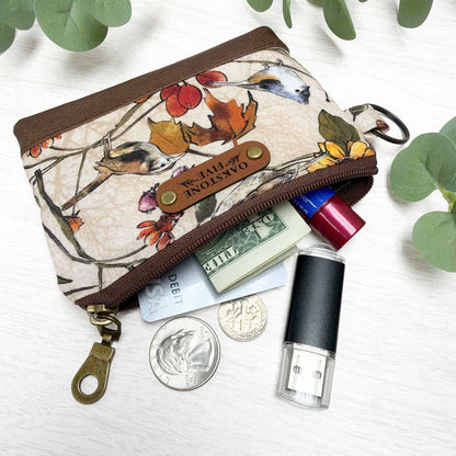 A cotton zippered coin purse with a brown and beige autumn bird print, shown open with various items inside on a white wood background with green leaves.