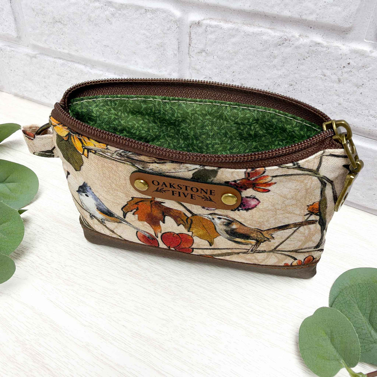 A small zippered pouch with a beige floral and bird print exterior, a green vines fabric interior, and a brown faux leather "Oakstone Five" brand tag. It's photographed on a white surface with green leaves in the foreground and a white brick wall behind.