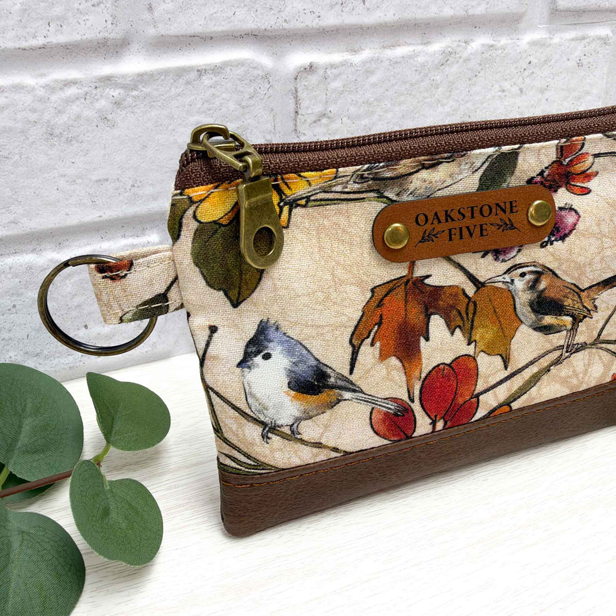 A fabric zip pouch with a brown faux-leather bottom, featuring a printed autumn leaf and bird pattern. It has a brown faux leather "Oakstone Five" brand tag and an antique brass zipper pull and key ring.
