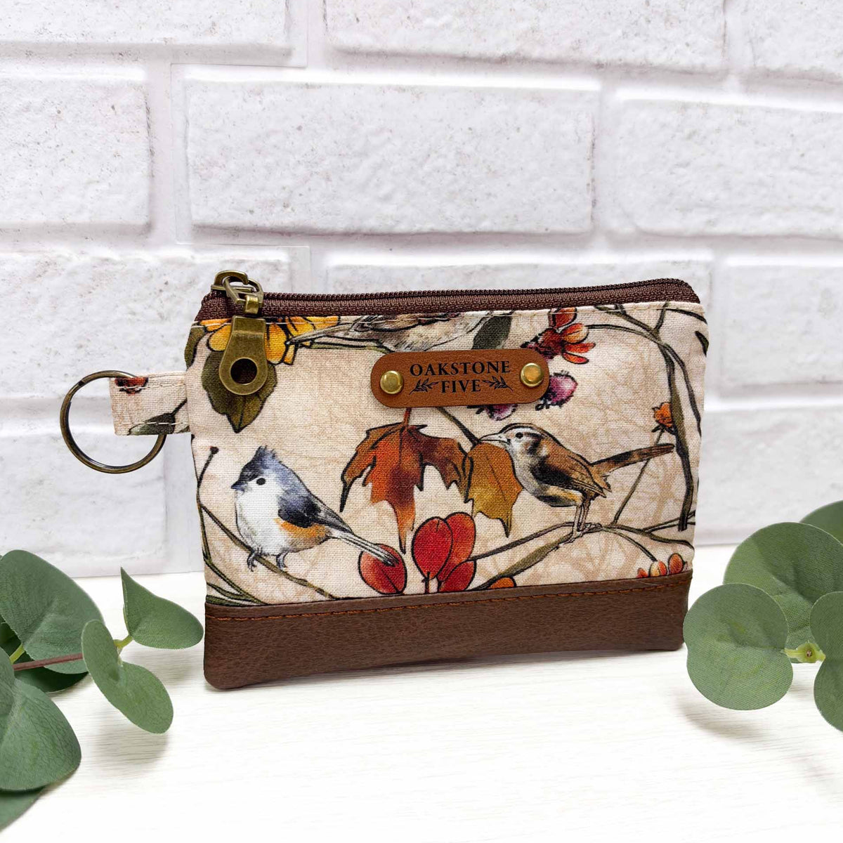 A small zip pouch with a cream background and a detailed bird and autumn leaf print, featuring brown faux leather accent, a brass key ring, and a faux leather "Oakstone Five" brand tag.