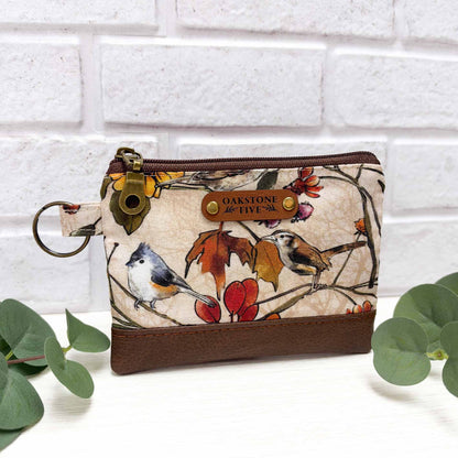 A small zip pouch with a cream background and a detailed bird and autumn leaf print, featuring brown faux leather accent, a brass key ring, and a faux leather "Oakstone Five" brand tag.