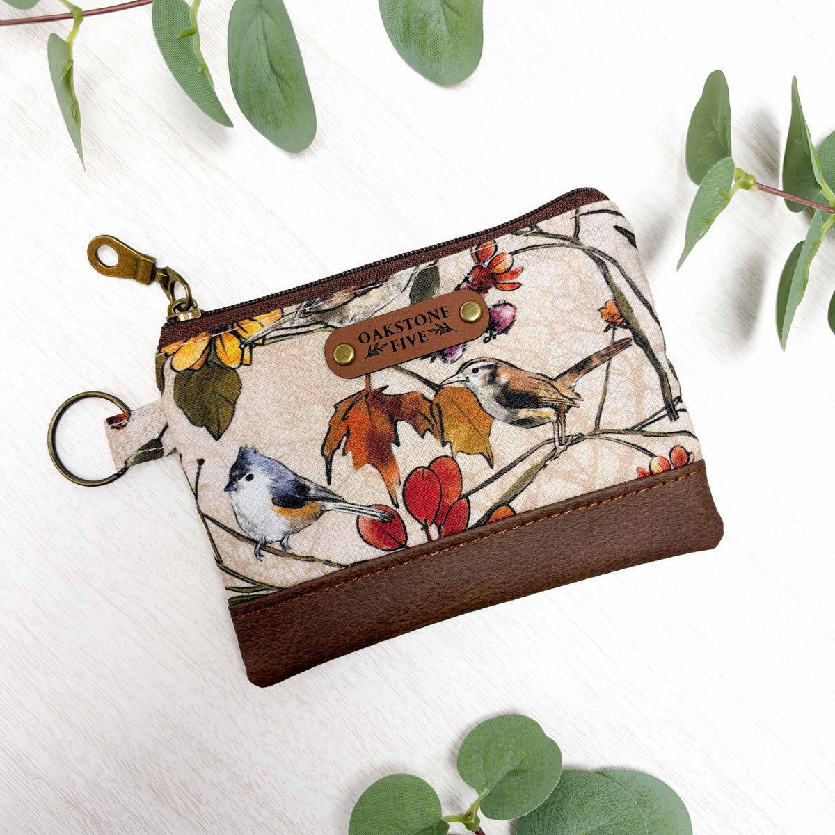 A small zip pouch with a cream background and a bird-and-branch print, featuring brown faux-leather bottom and  brass hardware. The pouch is photographed on a white surface with green leaves around it.