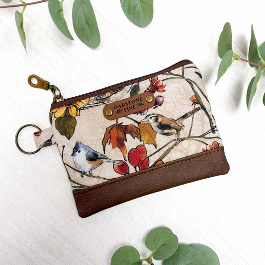 A small zip pouch with a cream background and a bird-and-branch print, featuring brown faux-leather bottom and  brass hardware. The pouch is photographed on a white surface with green leaves around it.