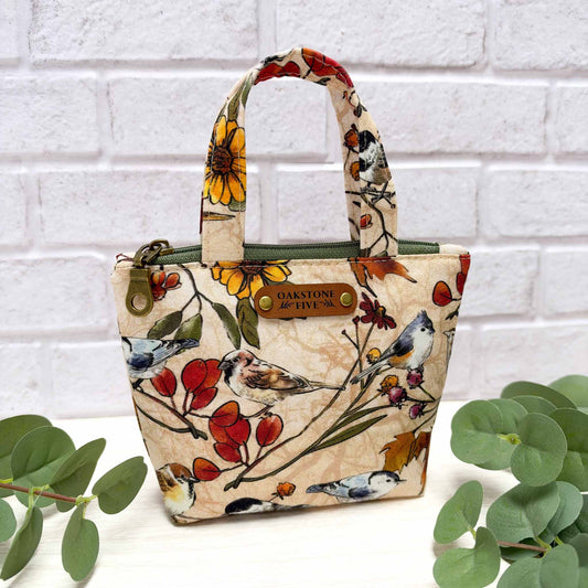 A mini fabric tote bag with a floral and bird print, featuring two top handles, a top zipper, and a brown faux leather "Oakstone Five" brand tag. The bag is styled on a white surface with green eucalyptus leaves on either side, set against a white brick wall background.