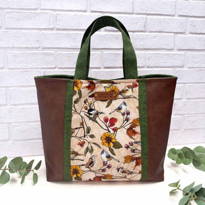 A tote bag with brown faux-leather sides, green fabric trim, a central printed panel featuring birds and autumn flowers on a beige background, with a small faux leather Oakstone Five brand patch on the front. 