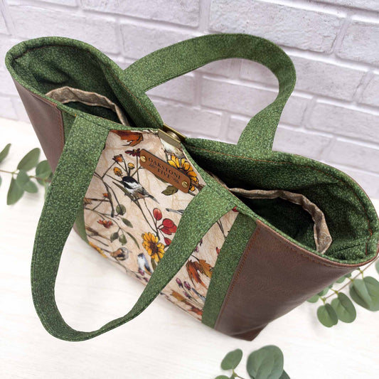 A tote bag with olive green fabric handles and side panels, brown faux leather side panels, and a floral and bird printed front panel, photographed on a white surface with a white brick wall background and small green leaves around it.