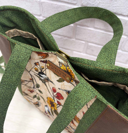 A bird lovers, nature-inspired tote bag with brown faux leather side panels, and green vines fabric handles and interior, photographed against a white brick wall background.