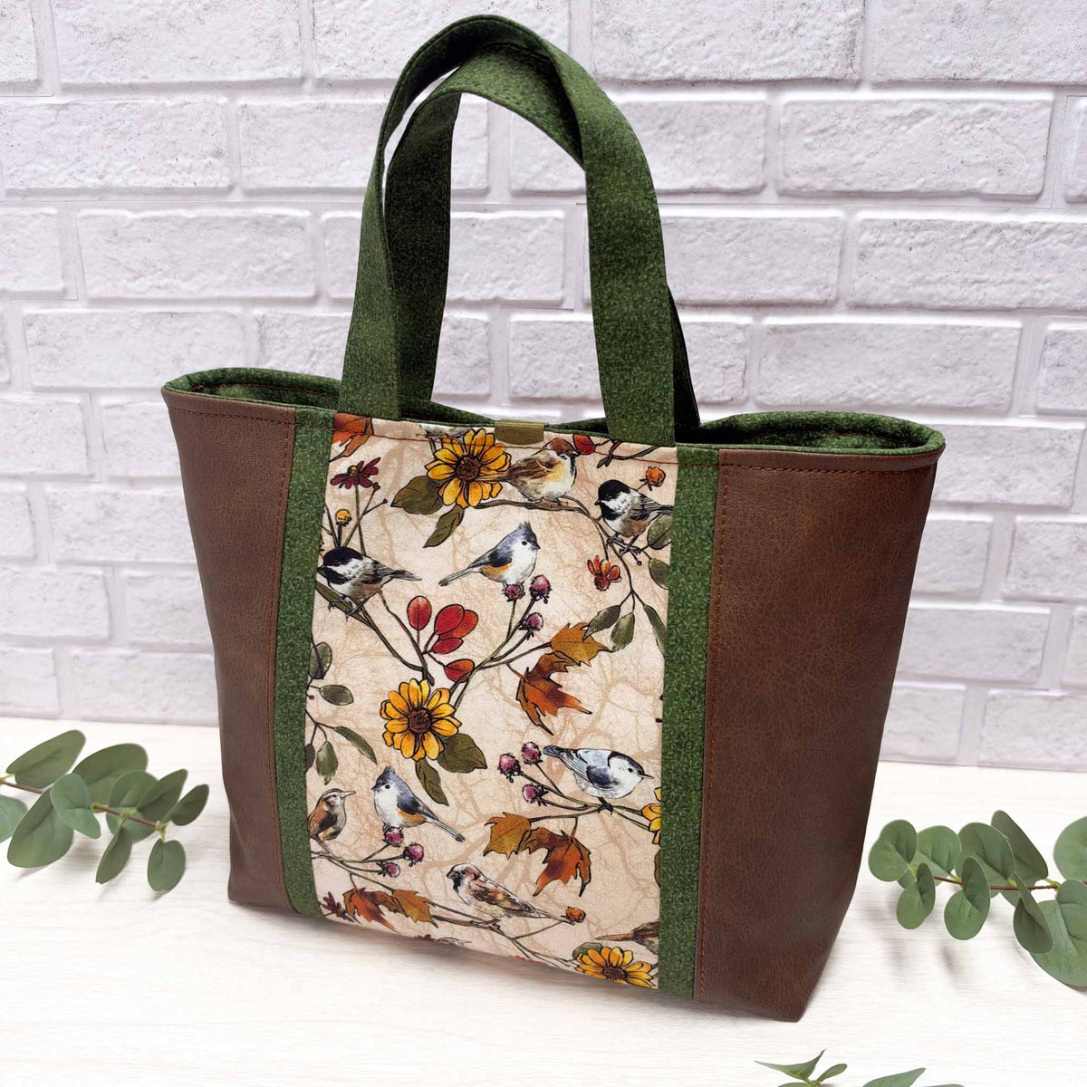 The back side of a tote bag with brown faux-leather sides, featuring a central panel with a bird and floral print on a beige background, set against a white brick wall backdrop.
