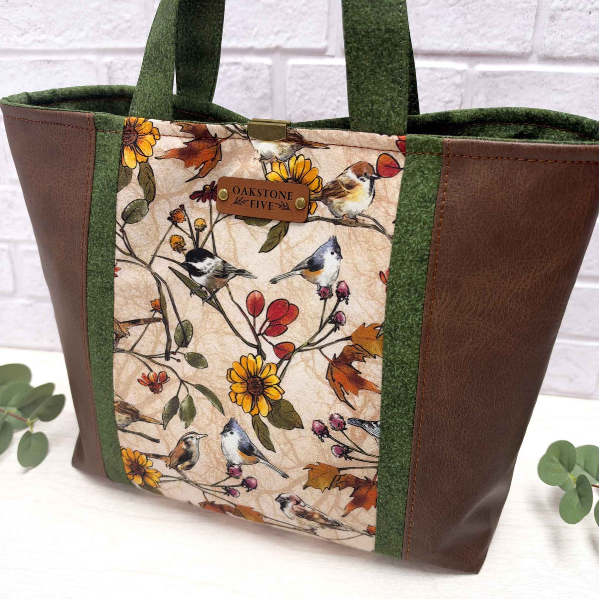 A tote bag with brown faux-leather sides, green fabric side panels, a central printed panel featuring birds and autumn flowers on a beige background, with a faux leather brand Oakstone Five tag.