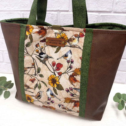 A tote bag with brown faux-leather sides, green fabric side panels, a central printed panel featuring birds and autumn flowers on a beige background, with a faux leather brand Oakstone Five tag.
