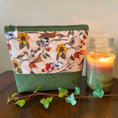 Large zipper pouch with flower and bird pattern, sitting next to a lit candle on a wooden surface with some ivy