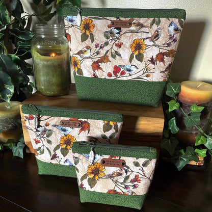 Set of three fabric zipper pouches with floral and bird pattern on a wooden surface with candles and plants.