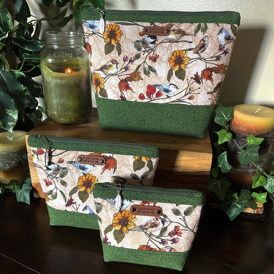 Set of three fabric zipper pouches with floral and bird pattern on a wooden surface with candles and plants.