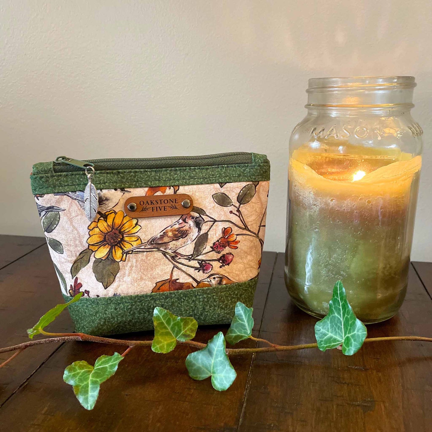 Small zipper pouch with flower and bird pattern, sitting next to a lit candle on a wooden surface with some ivy