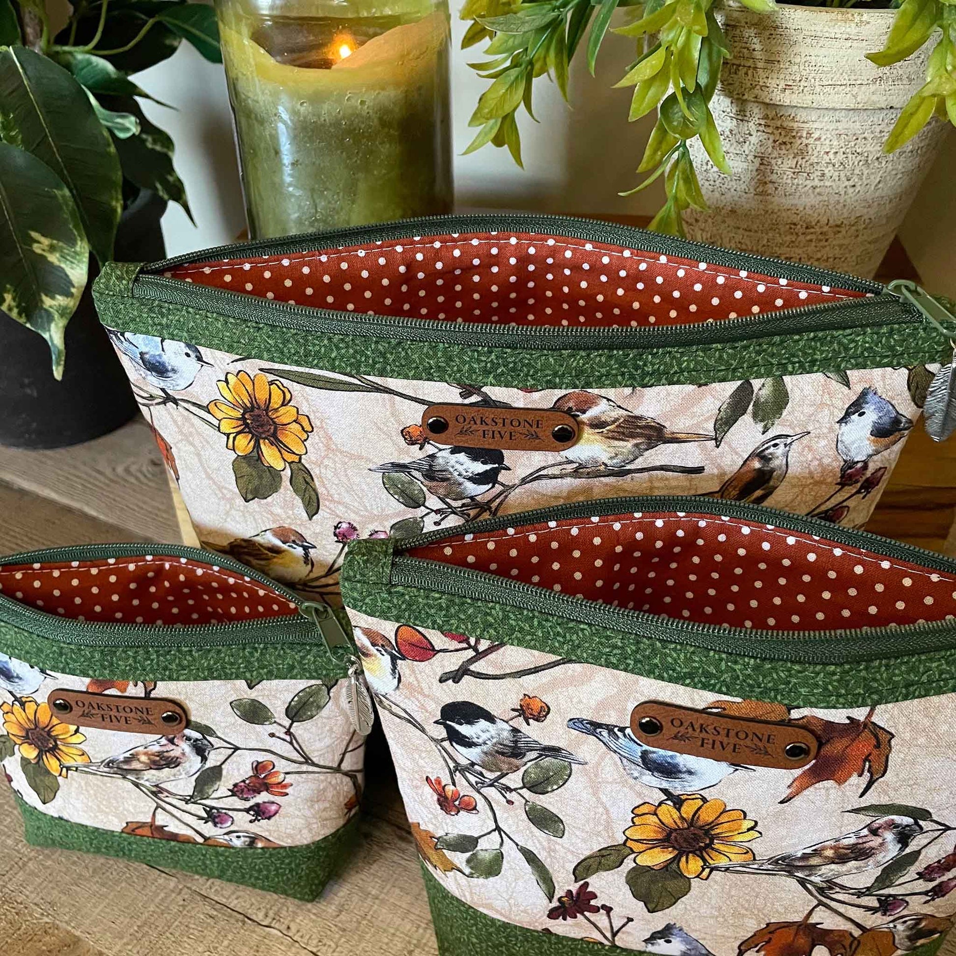 Three nature-themed zipper pouches with a birds and flower pattern and  an orange polka dot interior, sitting on a table with plants and candle in the background.