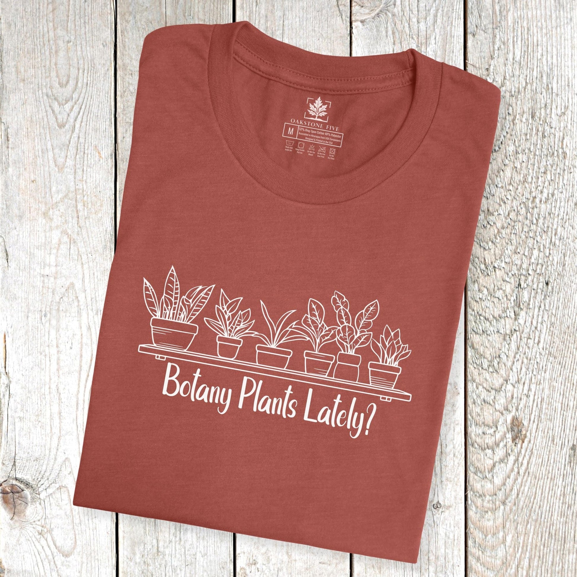 A folded, clay-colored, short-sleeve, funny plant t-shirt for gardeners and botanists that has a design with potted plants and says, "Botany Plants Lately?"