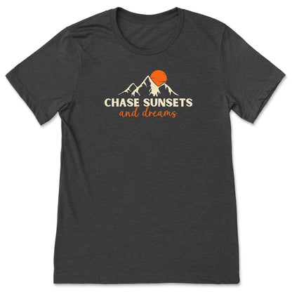 A dark grey, chasing sunsets t-shirt that has mountains with an orange sunset and says, "Chase Sunsets and Dreams."