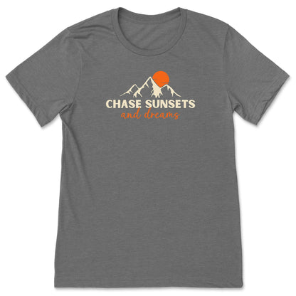 A grey chasing sunsets t-shirt that has mountains with an orange sunset and says, "Chase Sunsets and Dreams."