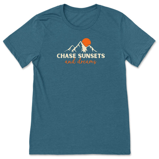 A teal chasing sunsets t-shirt that has mountains with an orange sunset and says, "Chase Sunsets and Dreams."