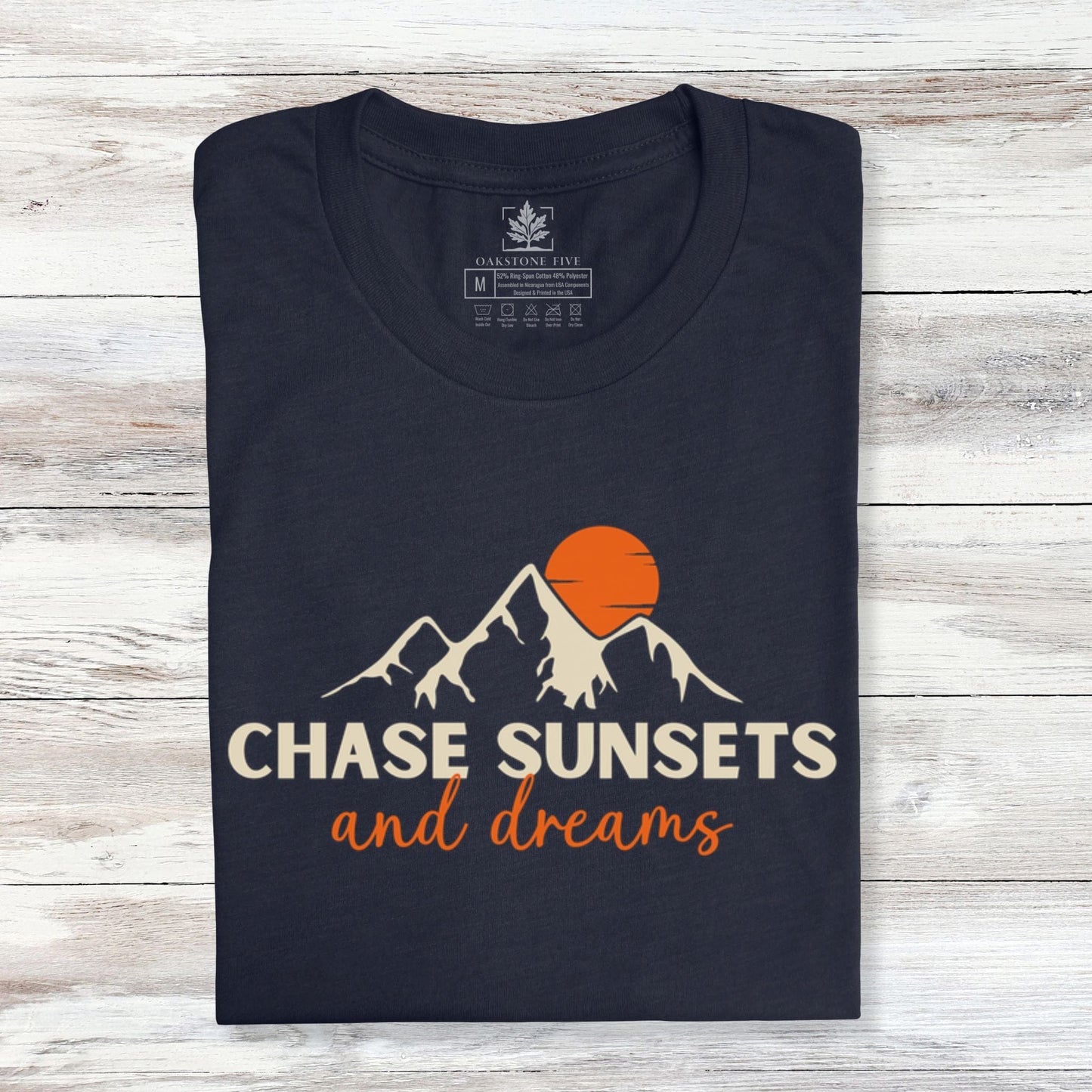 A folded, navy nature t-shirt that has mountains with an orange sunset and says, "Chase Sunsets and Dreams."