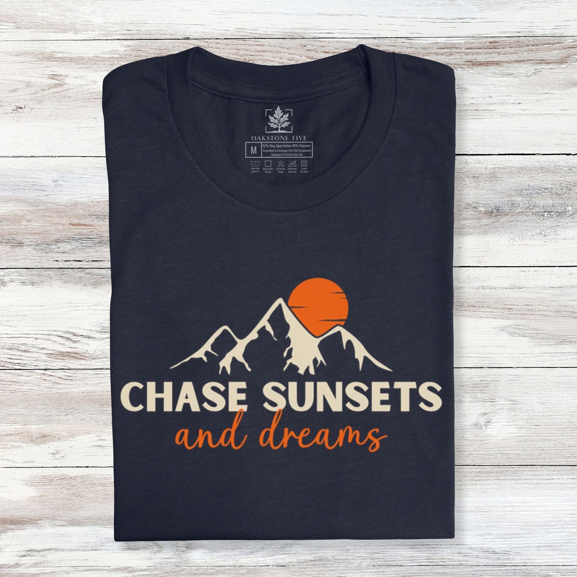 A folded, navy nature t-shirt that has mountains with an orange sunset and says, "Chase Sunsets and Dreams."