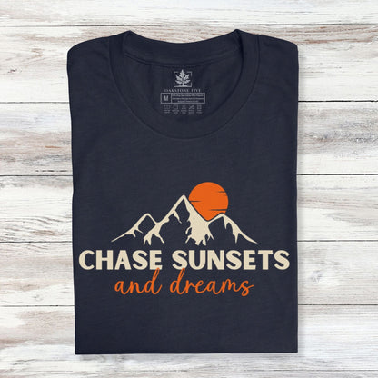 A folded, navy nature t-shirt that has mountains with an orange sunset and says, "Chase Sunsets and Dreams."