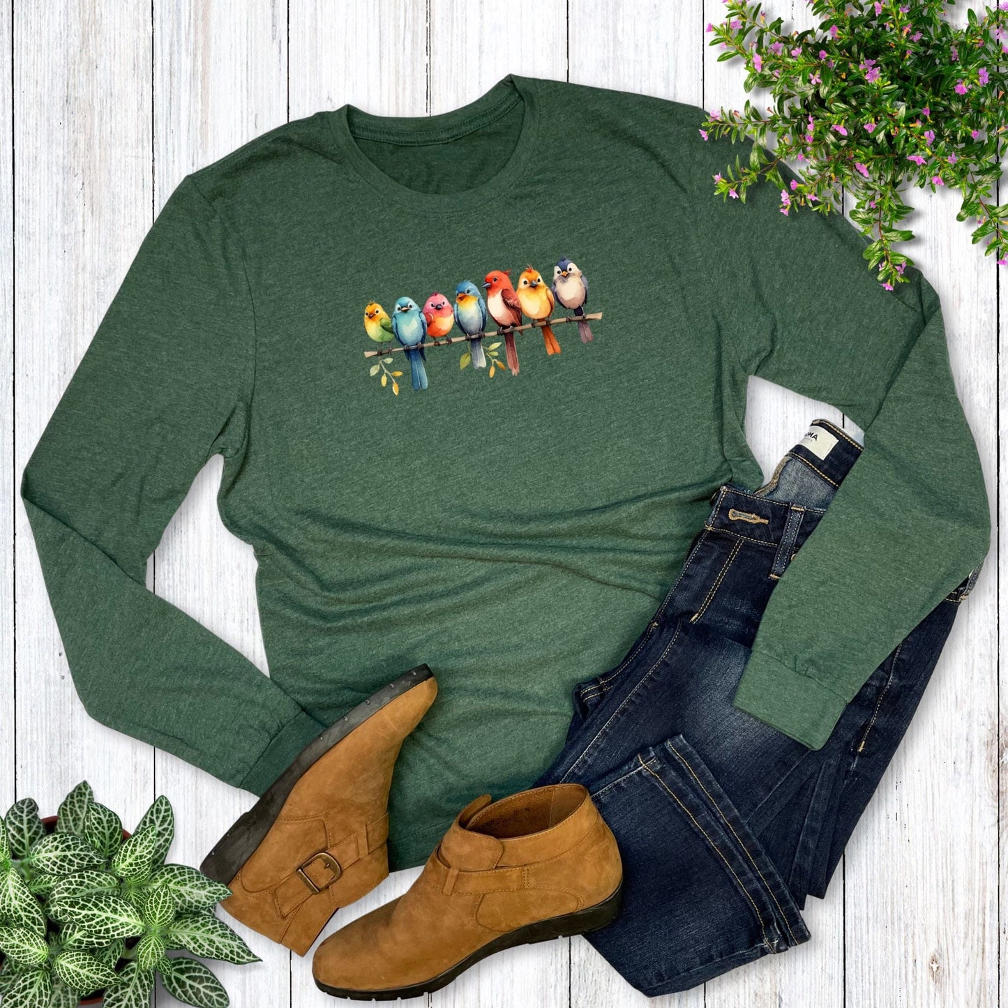 A long-sleeve, green t-shirt with a graphic of colorful birds sitting in a row on a branch, laying on a wooden surface with jeans, shoes, and plants.