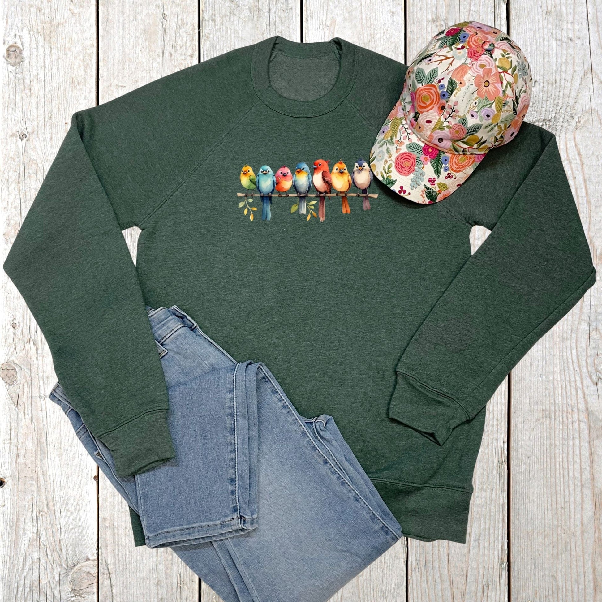 Green sweatshirt with colorful bird design, floral cap, and blue jeans on a wooden surface