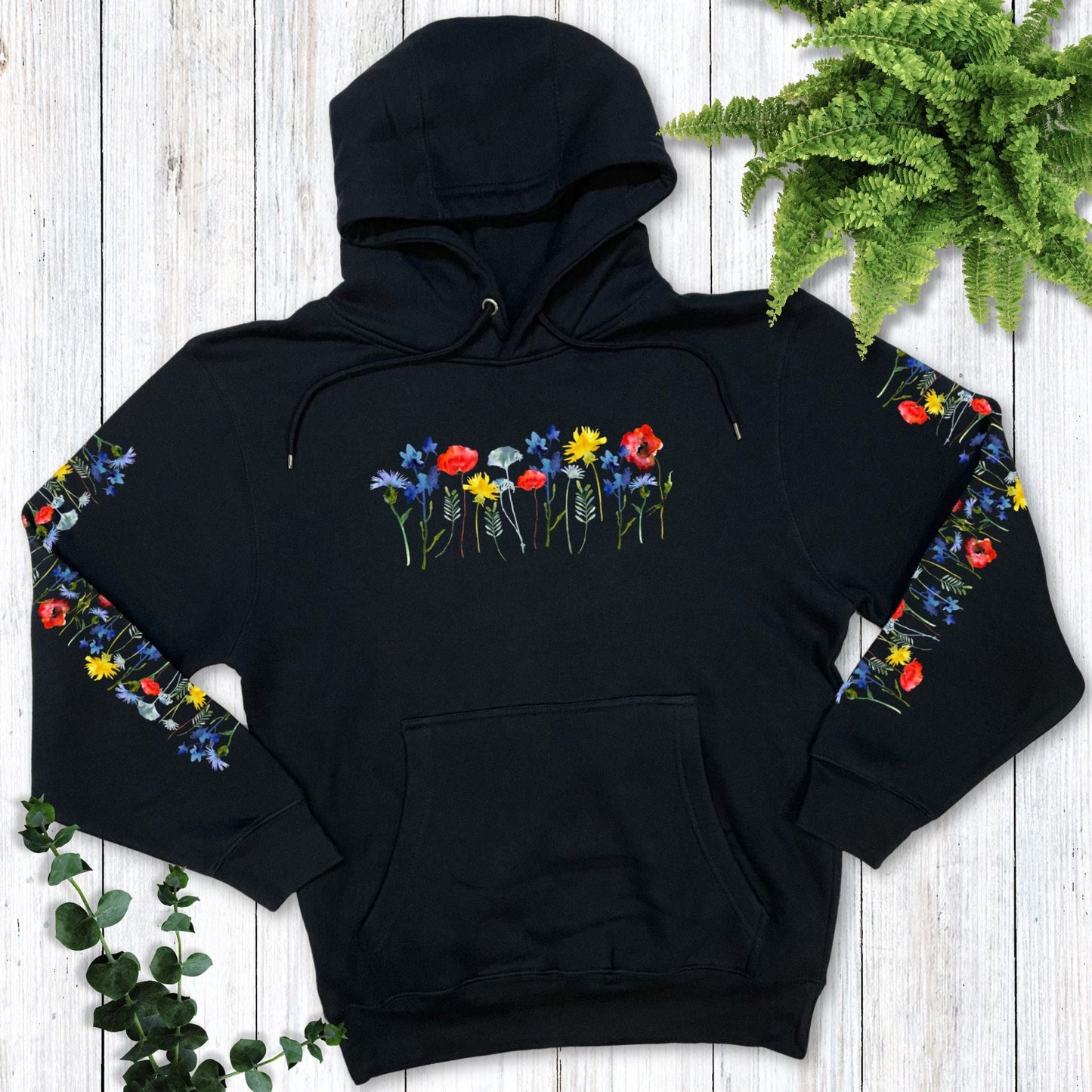 A navy hoodie for flower lovers that has vibrant, colorful flowers on the front and down both sleeves (and the back), laying on a wooden surface with greenery
