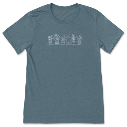 A cute, slate blue, plant t-shirt that has a simple line art drawing of potted plants on a shelf with a cat sitting between them - a great gift for plant lovers and gardeners!