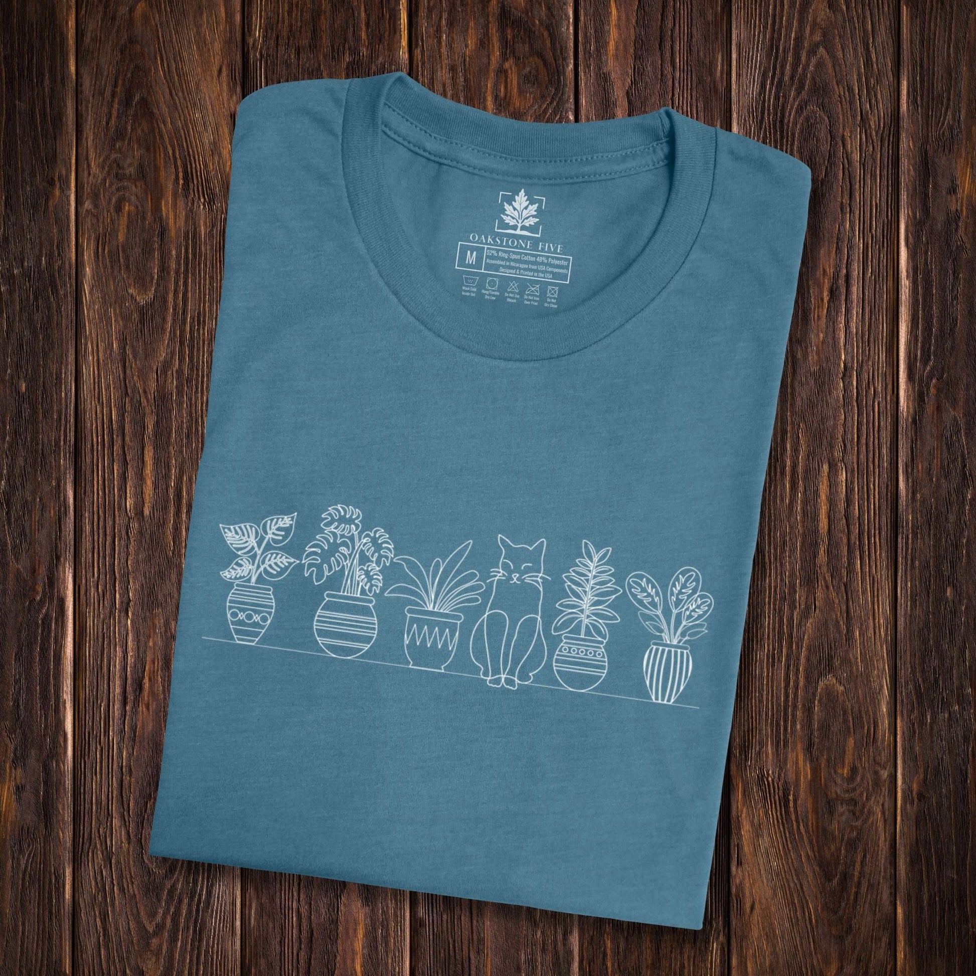 A folded cute, teal, plant t-shirt that has a simple line art drawing of potted plants on a shelf with a cat sitting between them - a great gift for plant lovers and gardeners!