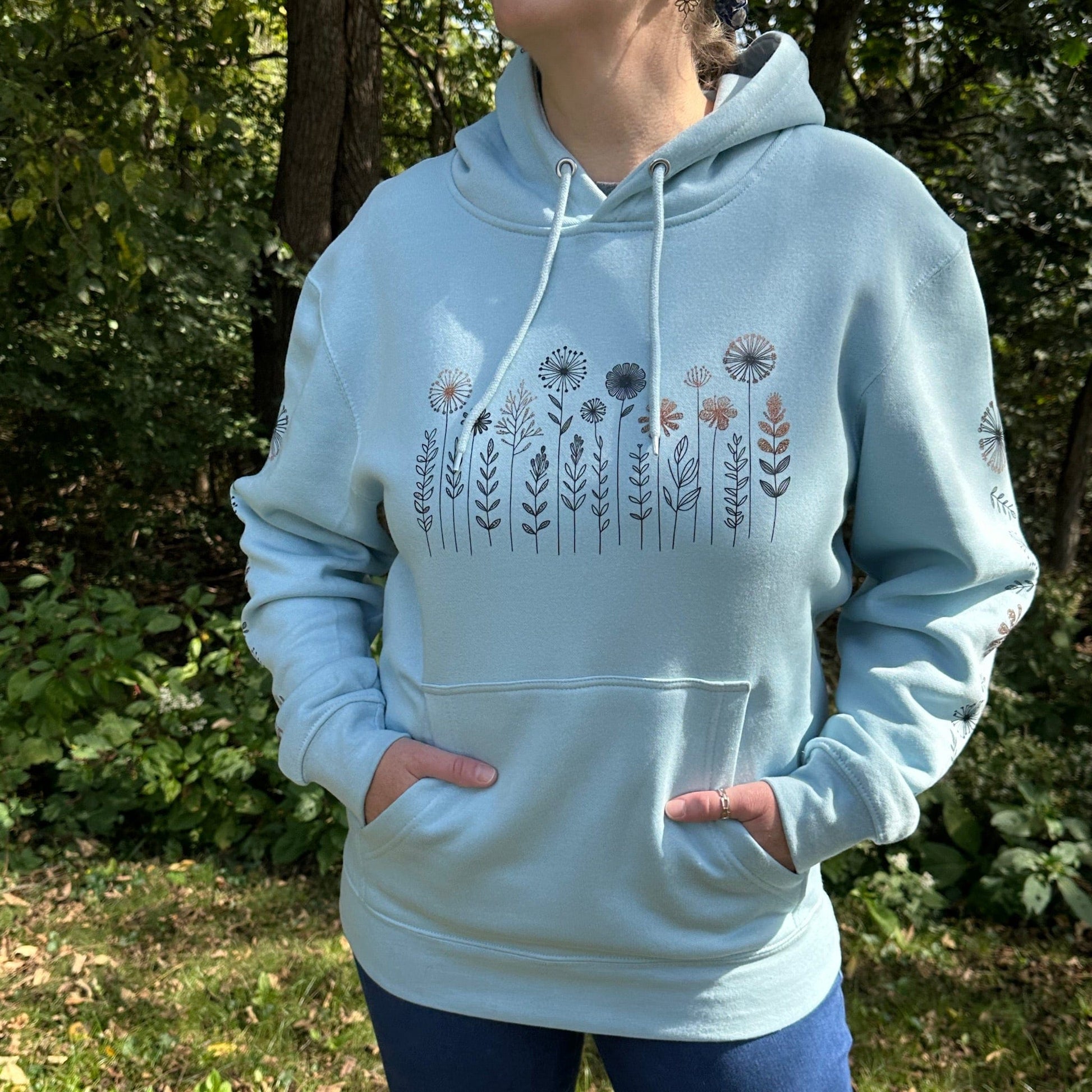 Woman standing in outside wearing a light blue hoodie that has a flower design on the chest and down both sleeves.