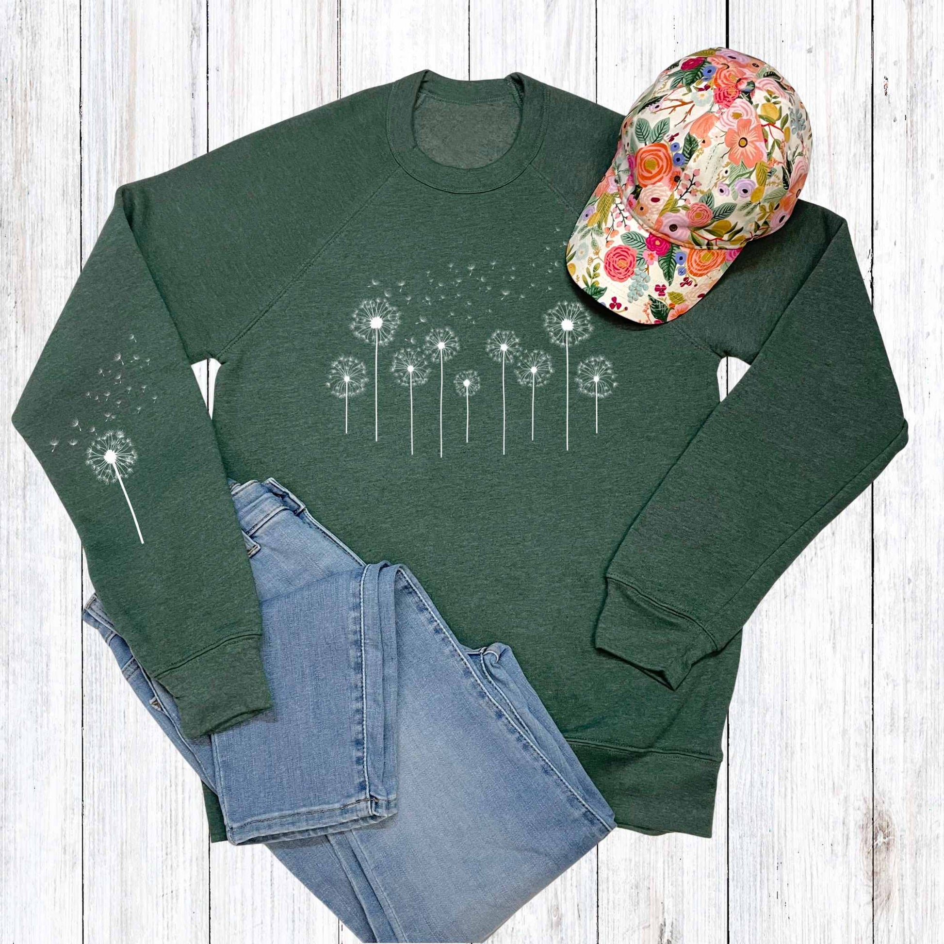 Green sweatshirt with dandelion design, floral cap, and blue jeans on a wooden surface