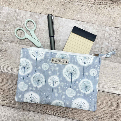 Fabric pouch with dandelion pattern on a wooden surface with stationery items.