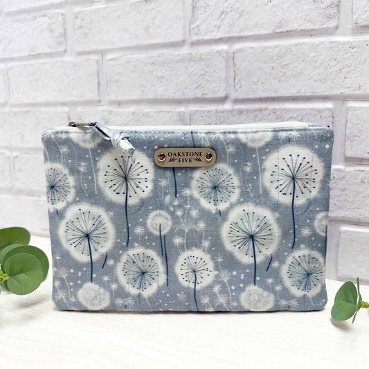 Quilted zipper pouch with blue and white dandelion pattern sitting on a white surface with leaves, against a white brick wall.