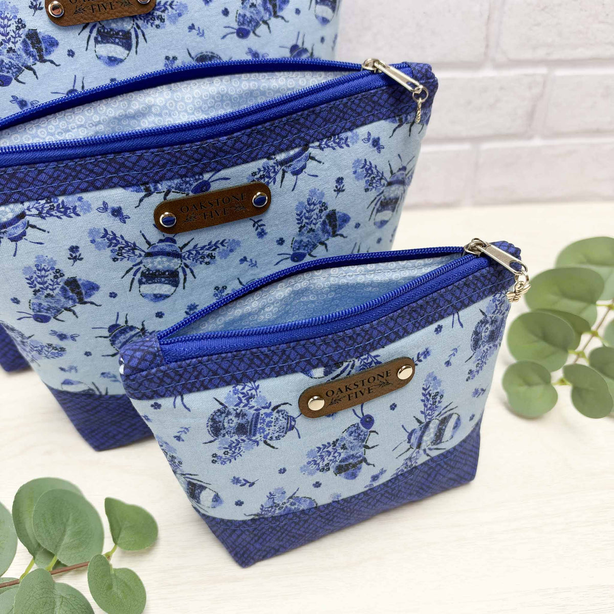 A close-up of three open zipper pouches with a blue floral bee pattern on the front with royal blue accents, and light blue patterned fabric on the inside, sitting on a white surface with leaves, in front of a white brick wall.