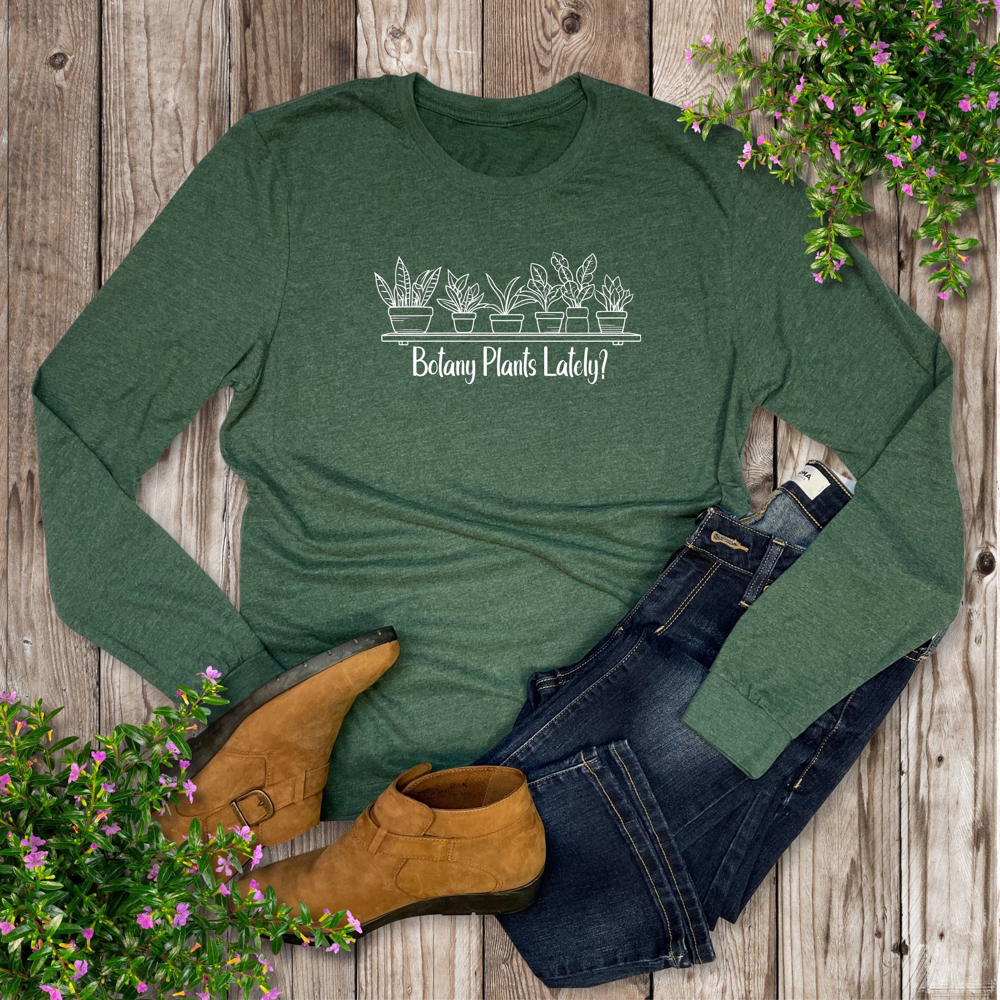 A green, funny botany long sleeve shirt for plant lovers that says, "Botany Plants Lately?", laying on a wooden surface surrounded by jeans, shoes, and plants.