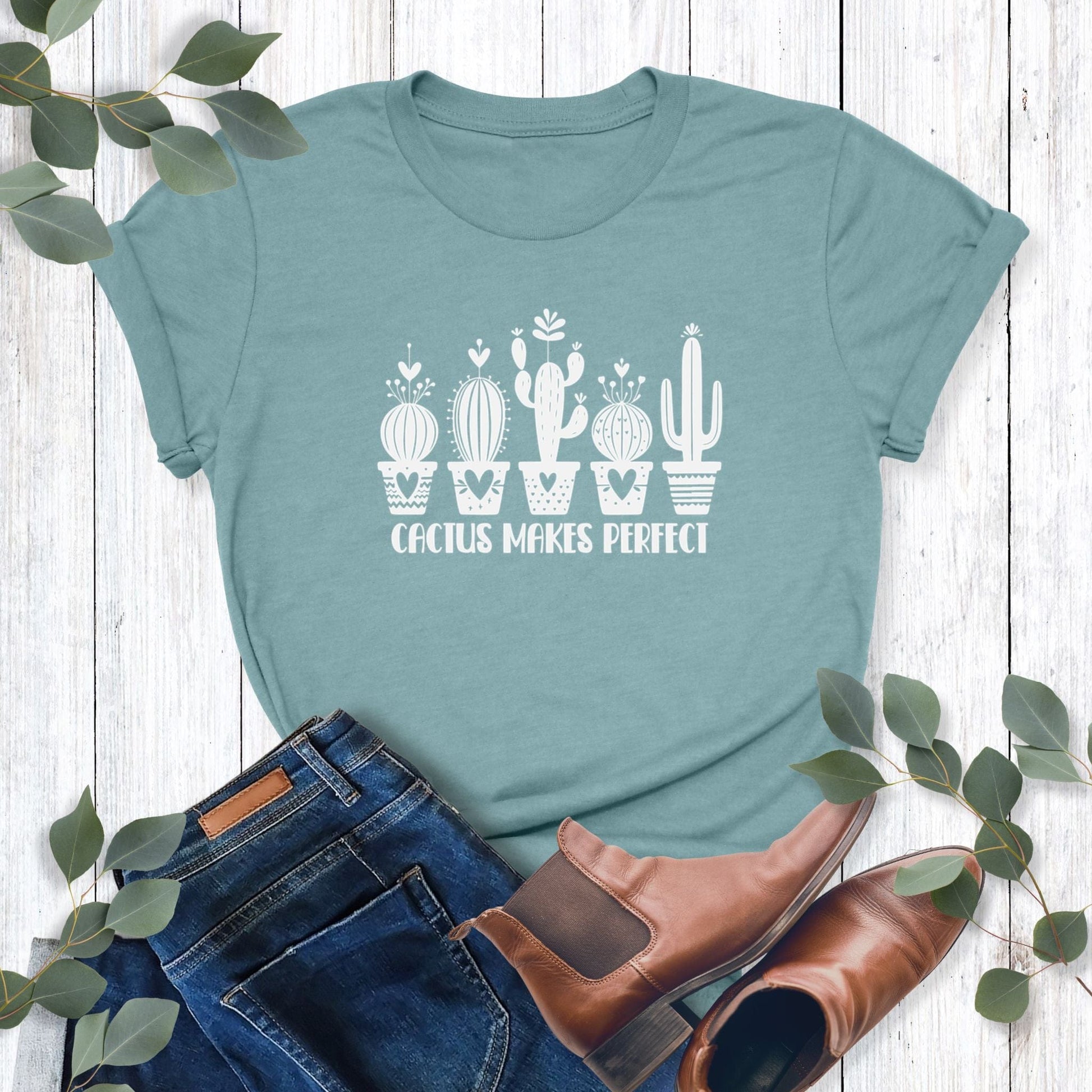 A light blue-green, funny plant t-shirt laying on a table with jeans and shoes.  Shirt design says, "Cactus Makes Perfect."