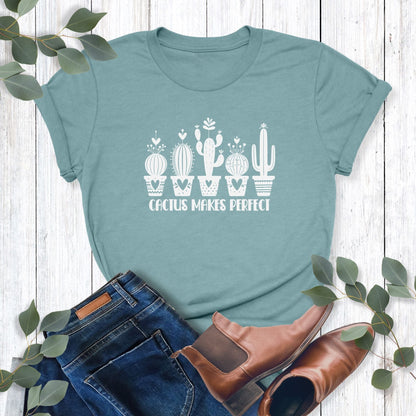 A light blue-green, funny plant t-shirt laying on a table with jeans and shoes.  Shirt design says, "Cactus Makes Perfect."