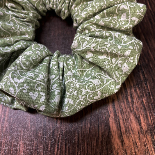 A close-up of a green scrunchie with white floral and bird pattern on a wood background