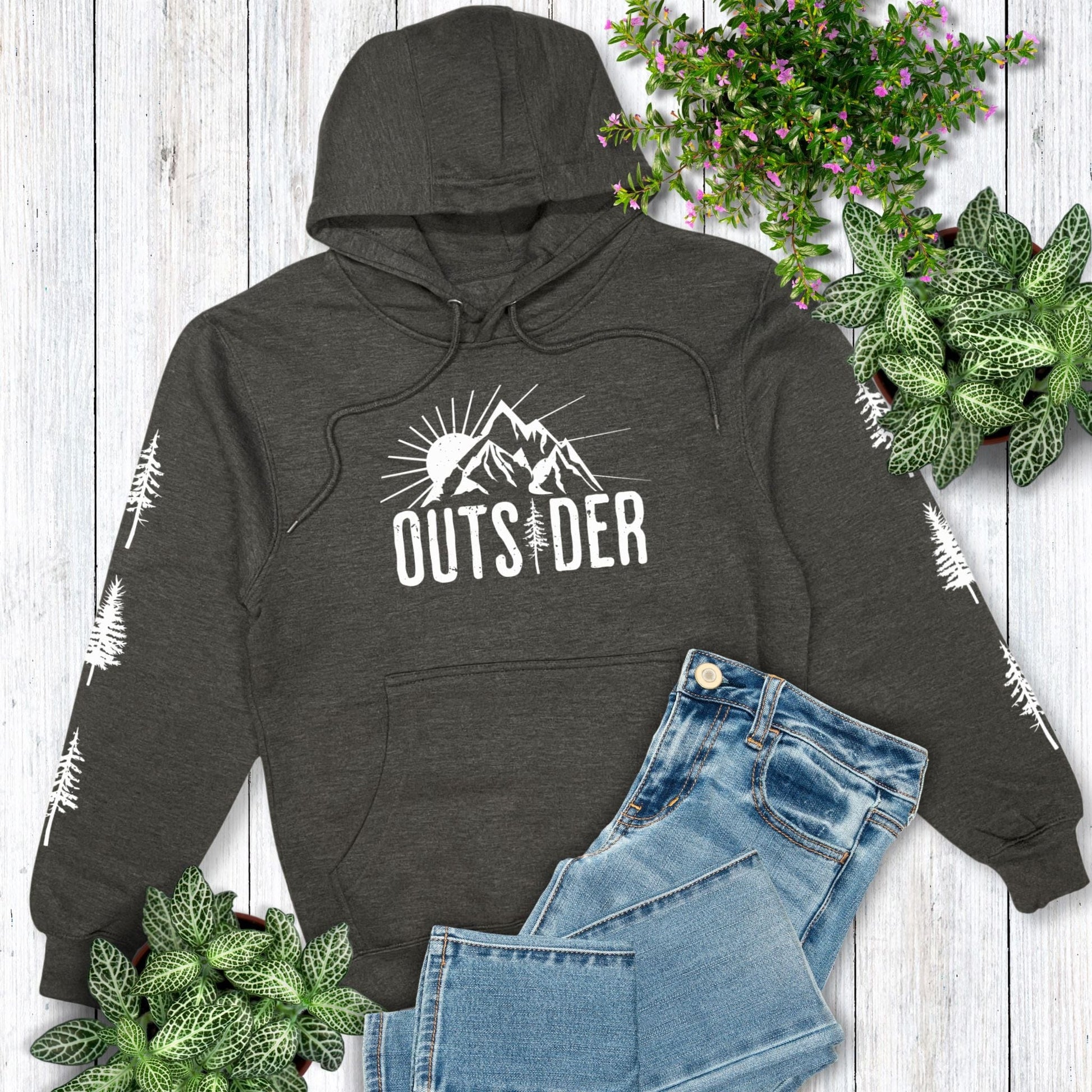 A dark grey hoodie for nature lovers that says "Outsider" on the front and has trees down both sleeves, laying on a wooden surface with jean and greenery